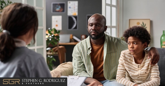 A man and his child sit in a couch and listen to a woman with a clipboard.