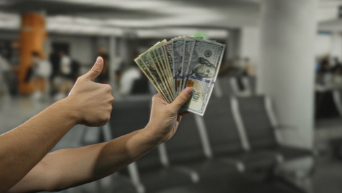Man holding american dollars inside airport with arm extended to show money and thumb up gesture amid empty waiting seats, symbolizing travel finance and optimism.